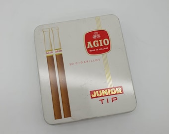 Può includere: Una scatola di metallo bianca con un'etichetta rossa e dorata. L'etichetta dice "Agio Junior Tip" e "20 Cigarillos" con un logo rosso e bianco di una corona e la parola "Agio" al centro. La scatola contiene due sigarette marroni.