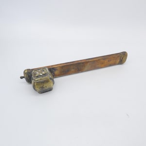 May include: A vintage brass and copper pipe with an ornate square chamber and a long, rectangular stem. The metal has a tarnished, aged appearance, with detailed carvings on the chamber.