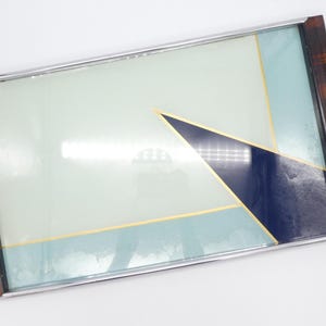 以下が含まれることがあります： 幾何学模様の長方形の装飾トレイ。トレイは、ライトブルーとネイビーブルーのパターンにゴールドのアクセントが施されています。フレームは濃い茶色で、シルバーのトリムが付いています。モダンなインテリアです。
