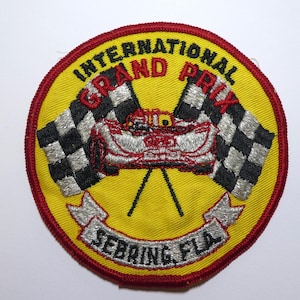 Può includere: Una toppa ricamata gialla con bordo rosso. La toppa presenta una bandiera a scacchi, un'auto da corsa e il testo "International Grand Prix Sebring FLA".