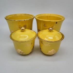 Peut inclure: Quatre pots de cuisine en céramique jaune avec des garnitures et des couvercles dorés. Deux grands pots ouverts et deux petits pots avec couvercles sont visibles. Les petits pots portent les mots "Rape" et "Droghe" en script doré. Les grands pots portent les mots "Sale" et "Küche".