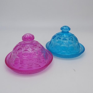 May include: Two glass butter dishes, one magenta and one turquoise. Each dish has a domed lid with a knob and a circular base. The lids and bases have a textured, patterned design. The dishes are on a white background.