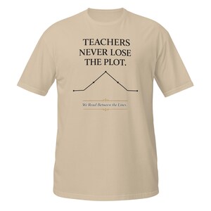 May include: A beige short-sleeve t-shirt featuring the black text TEACHERS NEVER LOSE THE PLOT above a black line graph illustration. Below the graph, the text We Read Between the Lines is printed in a smaller font with decorative gold accents.