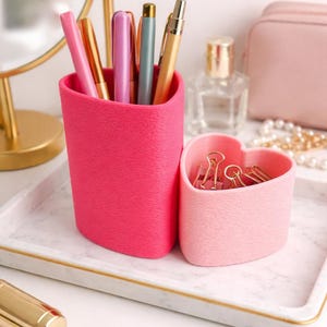 May include: A vibrant pink pen holder and a heart-shaped container, both with textured surfaces. The pen holder contains pens, and the heart-shaped container holds paper clips. They sit on a marble tray with a gold trim, suitable for a desk or dressing table.