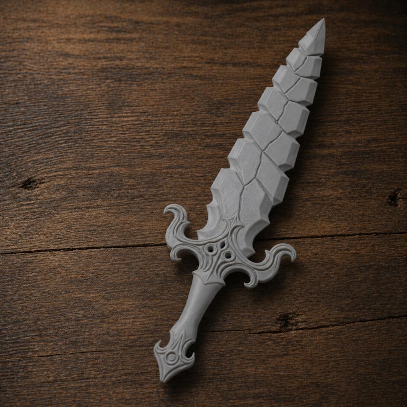 May include: A gray decorative sword with a detailed handle and a textured blade. The blade has a segmented design, and the handle features ornate swirls and cutouts. The sword is displayed on a wooden surface.