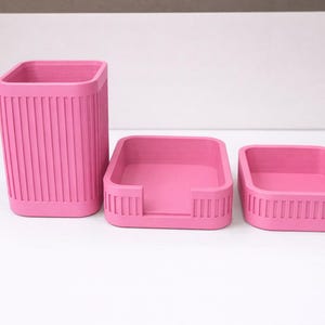 Könnte beinhalten: Ein Set aus drei passenden rosa Schreibtisch-Organizern. Das Set beinhaltet einen hohen, rechteckigen Stifthalter und zwei kleinere, quadratische Tabletts. Alle Teile haben ein geripptes Design.