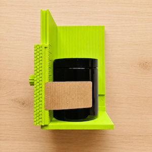 May include: A bright lime green plastic centering tool with a textured honeycomb side panel and integrated ruler markings. A cylindrical black jar with a smooth finish is positioned in the center, featuring a rectangular tan cardboard label.
