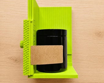 May include: A bright lime green plastic centering tool with a textured honeycomb side panel and integrated ruler markings. A cylindrical black jar with a smooth finish is positioned in the center, featuring a rectangular tan cardboard label.