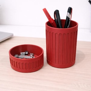 May include: Red desk organizer set with a pen holder and a small round container. The pen holder contains pens and pencils. The small container holds small metal objects. The set is on a light wood surface.
