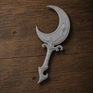May include: A gray, crescent-shaped decorative object with ornate detailing. The design features a stylized leaf and swirling patterns on the crescent and handle. The object is made of a solid material and rests on a wooden surface.