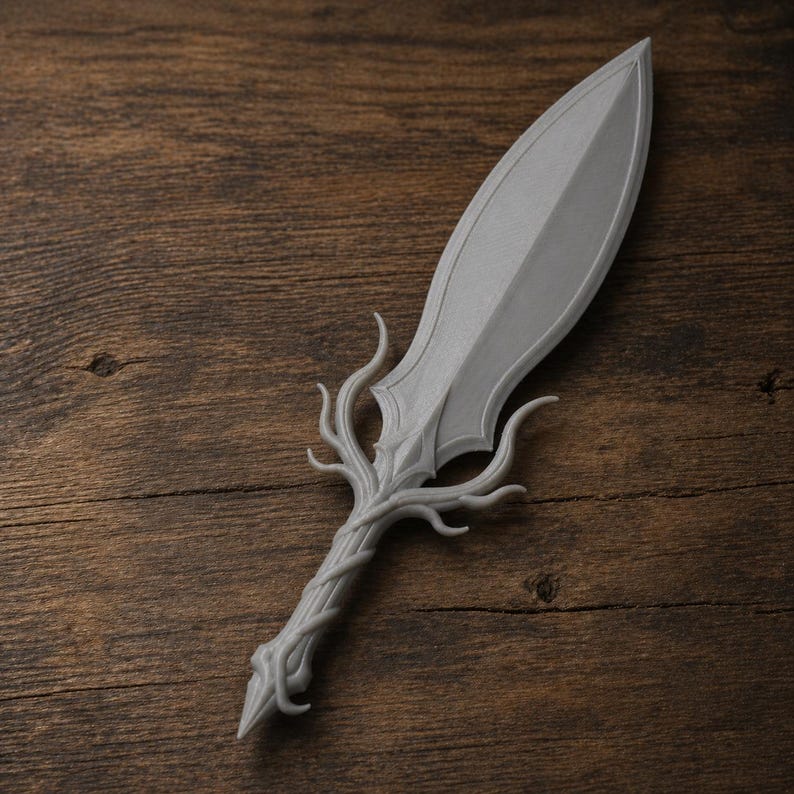 May include: A gray, decorative dagger with a leaf-shaped blade and a handle designed to resemble intertwined branches. The dagger is made of a solid material and rests on a wooden surface.