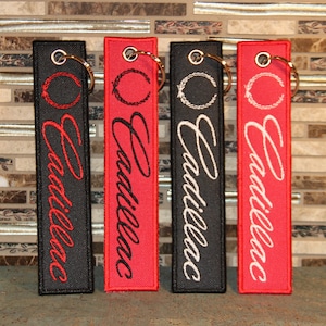 May include: Four keychains with a black, red, white, and red background. Each keychain has a white embroidered wreath and the word "Cadillac" in cursive.