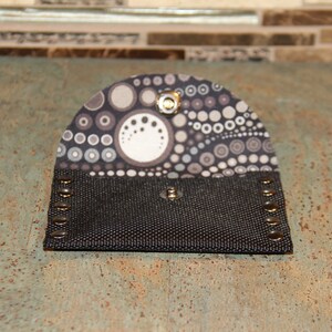 Pode incluir: Uma pequena carteira aberta com um corpo de lona preta e uma aba estampada. A aba apresenta um design de c&iacute;rculos e pontos cinza e brancos. A carteira &eacute; presa com um fecho de press&atilde;o e tem rebites de metal nas bordas.