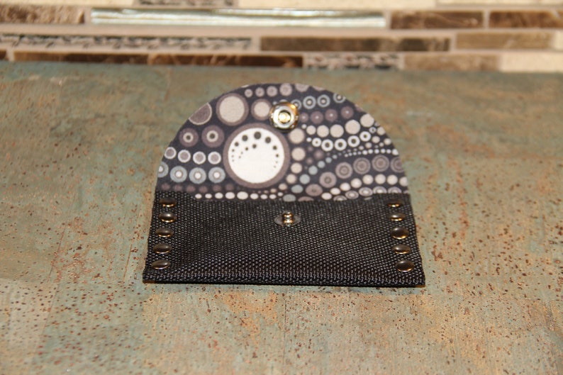 Pode incluir: Uma pequena carteira aberta com um corpo de lona preta e uma aba estampada. A aba apresenta um design de c&iacute;rculos e pontos cinza e brancos. A carteira &eacute; presa com um fecho de press&atilde;o e tem rebites de metal nas bordas.