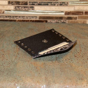 Pode incluir: Uma bainha preta em forma de diamante com um padr&atilde;o texturizado e rebites prateados nas bordas. A bainha tem um bot&atilde;o prateado no centro e uma borda decorativa. O item &eacute; exibido em uma superf&iacute;cie azul-esverdeada salpicada.