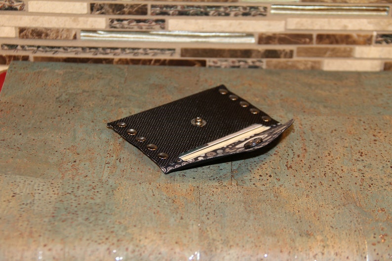 Pode incluir: Uma bainha preta em forma de diamante com um padr&atilde;o texturizado e rebites prateados nas bordas. A bainha tem um bot&atilde;o prateado no centro e uma borda decorativa. O item &eacute; exibido em uma superf&iacute;cie azul-esverdeada salpicada.