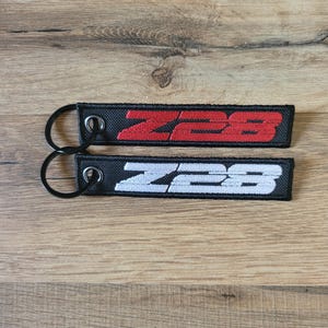 May include: Two black keychains with a white and red embroidered "Z28" logo. The keychains are on a wooden surface.