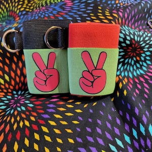 May include: Two black and green fabric sleeves with a pink peace sign on each. The sleeves have a red top and a black strap with a metal ring.