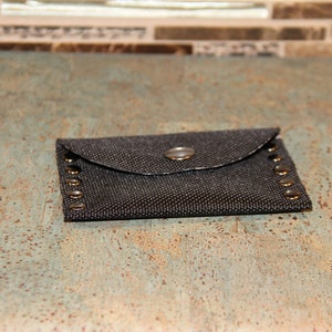 Pode incluir: Uma pequena carteira de tecido cinza escuro com fecho de aba e um &uacute;nico bot&atilde;o de press&atilde;o. A carteira &eacute; adornada com pequenos rebites redondos de cor bronze ao longo das bordas. A carteira tem aproximadamente 10 cm de comprimento e 6 cm de largura.