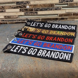May include: Five black keychains with white text that reads "Let's Go Brandon!" The keychains are stacked on top of each other. The top keychain is black with white text, the second keychain is blue with red text, the third keychain is red with white text, the fourth keychain is camouflage with yellow text, and the bottom keychain is brown with yellow text.