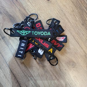 May include: A collection of black embroidered keychains with various car brand logos and text, including "TOYODA", "LEXUS", and "EVV". Each keychain has a metal ring for attachment. The keychains are arranged on a wooden surface.