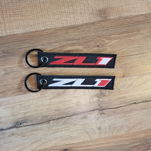 May include: Two black keychains with a white and red embroidered "ZL1" logo. The keychains are on a wooden surface.