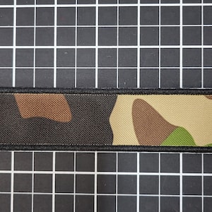 May include: A camouflage-patterned key chain with a brass key ring. The key chain is made of fabric and has a black border.
