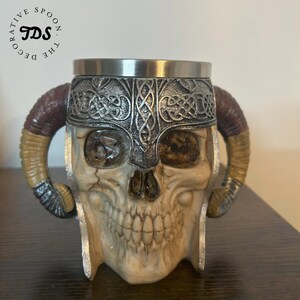 May include: A ceramic skull mug with a silver helmet and horns. The mug has a stainless steel lid and is decorated with a Celtic knot design.