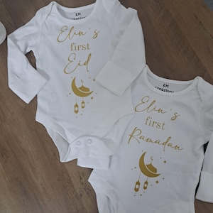 May include: Two white long-sleeved baby bodysuits featuring gold text and graphics. One reads 'Elin's first Eid' and the other 'Elin's first Ramadan'. Both include a gold crescent moon, a small dome, and hanging lantern illustrations with stars.