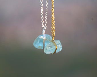 May include: Two necklaces with light blue gemstone pendants. One necklace has a silver chain, and the other has a gold chain. The gemstones are wrapped with wire and suspended from the chains.
