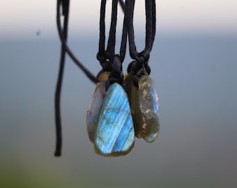 May include: Close-up of three labradorite stone pendants on black cords. The stones display iridescent blue and gold hues. The pendants are suspended against a blurred, neutral background, highlighting the natural beauty of the gemstones.