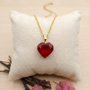 Puede incluir: Un collar con cadena dorada y colgante rojo en forma de corazón facetado. El collar se exhibe sobre un cojín cuadrado blanco roto. Ramitas secas y pétalos de flores rosas se disponen alrededor del cojín.