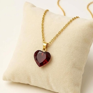Peut inclure: Un collier à chaîne dorée avec un pendentif en forme de cœur, facetté, rouge foncé. Le pendentif est suspendu à une simple bélière. Le collier est présenté sur une surface texturée de couleur crème.