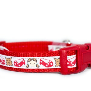 May include: Red dog collar with a white ribbon featuring a repeating pattern of a girl with pigtails, red shoes, and a dog. The collar has a red plastic buckle.