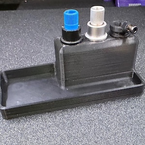 May include: A black 3D-printed device with a tray and three metallic components. The components include a blue cap, a silver cap, and a black fitting with a small nozzle. The device is designed for a specific function.