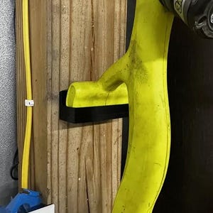 May include: A yellow plastic bow holder mounted on a wooden wall. The holder has a curved shape and a black base.