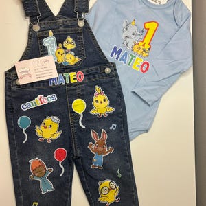 Op de afbeelding: Een set voor baby's bestaande uit een spijkerbroek en een lichtblauwe longsleeve romper. De spijkerbroek heeft stickers van stripfiguren en de naam "MATEO". De romper heeft een cartoon olifant en het nummer "1" erop gedrukt.