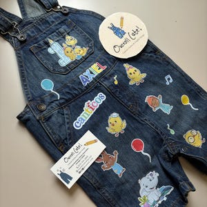 May include: Dark blue denim overalls featuring various colorful cartoon character stickers, including chicks, an elephant, and a rabbit. The text "AXIEL" and "Canticos" are printed on the front. Includes a round "Overall Cute!" brand tag.