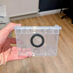 May include: A clear plastic terrarium with a mesh vent. The rectangular container has a hinged lid and is held by a hand. The vent is a black circle with a fine mesh screen. The background is a light-coloured wall and wooden floor.