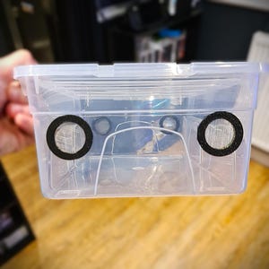 May include: A transparent plastic storage box with a lid, showing two circular mesh vents with black surrounds. The rectangular container is designed for storage. The image is taken from a side view.