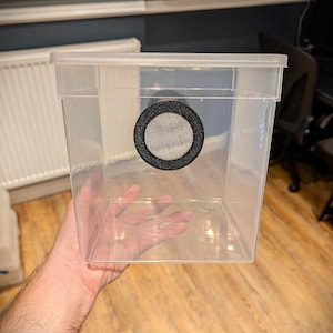May include: A clear, rectangular plastic container with a lid. A circular mesh vent is centered on the side. The container is held in a person's hand.
