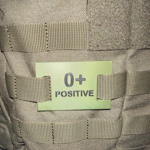 May include: A close-up of a tan tactical vest with a light green patch. The patch has the text "0+ POSITIVE" in black. The vest has multiple straps and a textured surface.