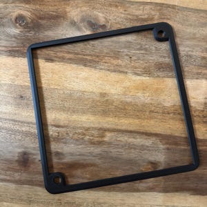 Op de afbeelding: Een zwart kentekenplaatraam met afgeronde hoeken en montagegaten. Het frame is vierkant en ligt op een houten oppervlak. Het frame is gemaakt van een stevig materiaal en heeft een matte afwerking.