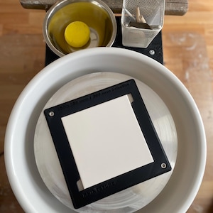 May include: A pottery wheel with a white ceramic bowl and a black frame holding a white square. A yellow ball sits in a metal bowl. The frame reads "300 x 300 mm / 300 mm Scheibe" and "PRINZTUBE".