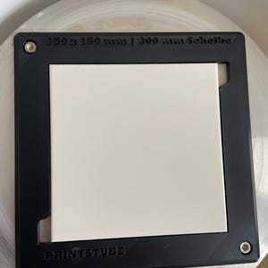 Pu&ograve; includere: Una cornice quadrata nera con un inserto quadrato bianco. La cornice presenta il testo "150 x 150 mm | 300 mm Scheibe" e "PRINTSTUBE". La cornice &egrave; su una superficie circolare di colore chiaro.