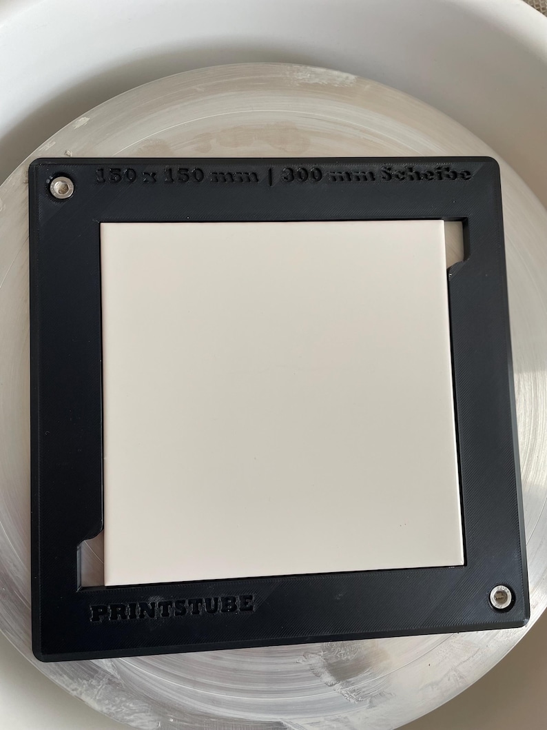 Pu&ograve; includere: Una cornice quadrata nera con un inserto quadrato bianco. La cornice presenta il testo "150 x 150 mm | 300 mm Scheibe" e "PRINTSTUBE". La cornice &egrave; su una superficie circolare di colore chiaro.