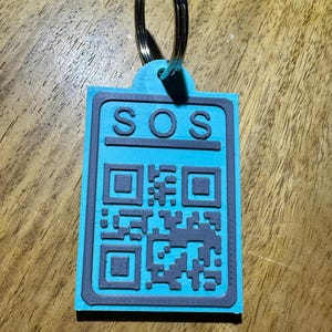 Könnte beinhalten: Ein türkisfarbener Schlüsselanhänger mit einem schwarz-weißen QR-Code und dem aufgedruckten Wort "SOS".