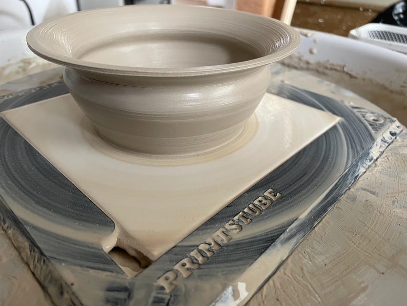 Pu&ograve; includere: Una ciotola in ceramica beige chiaro con un bordo largo, appoggiata su una superficie quadrata di colore chiaro. La ciotola ha una forma arrotondata ed &egrave; centrata su una piattaforma rotante. La parola "PRINTSTUBE" &egrave; visibile sulla piattaforma.