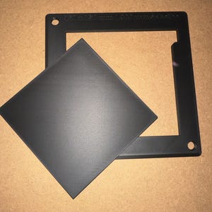 May include: A black square frame with a matching square insert. The frame has two holes and text along one side. The insert is slightly smaller than the frame. The items are on a light brown surface.
