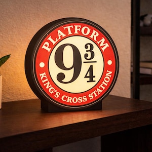 Peut inclure: Panneau rond et lumineux avec le texte "PLATFORM 9 3/4 KING'S CROSS STATION" en rouge et blanc. Le panneau est posé sur une surface marron foncé, avec une plante en pot à gauche. Une étagère avec des livres est visible en arrière-plan.